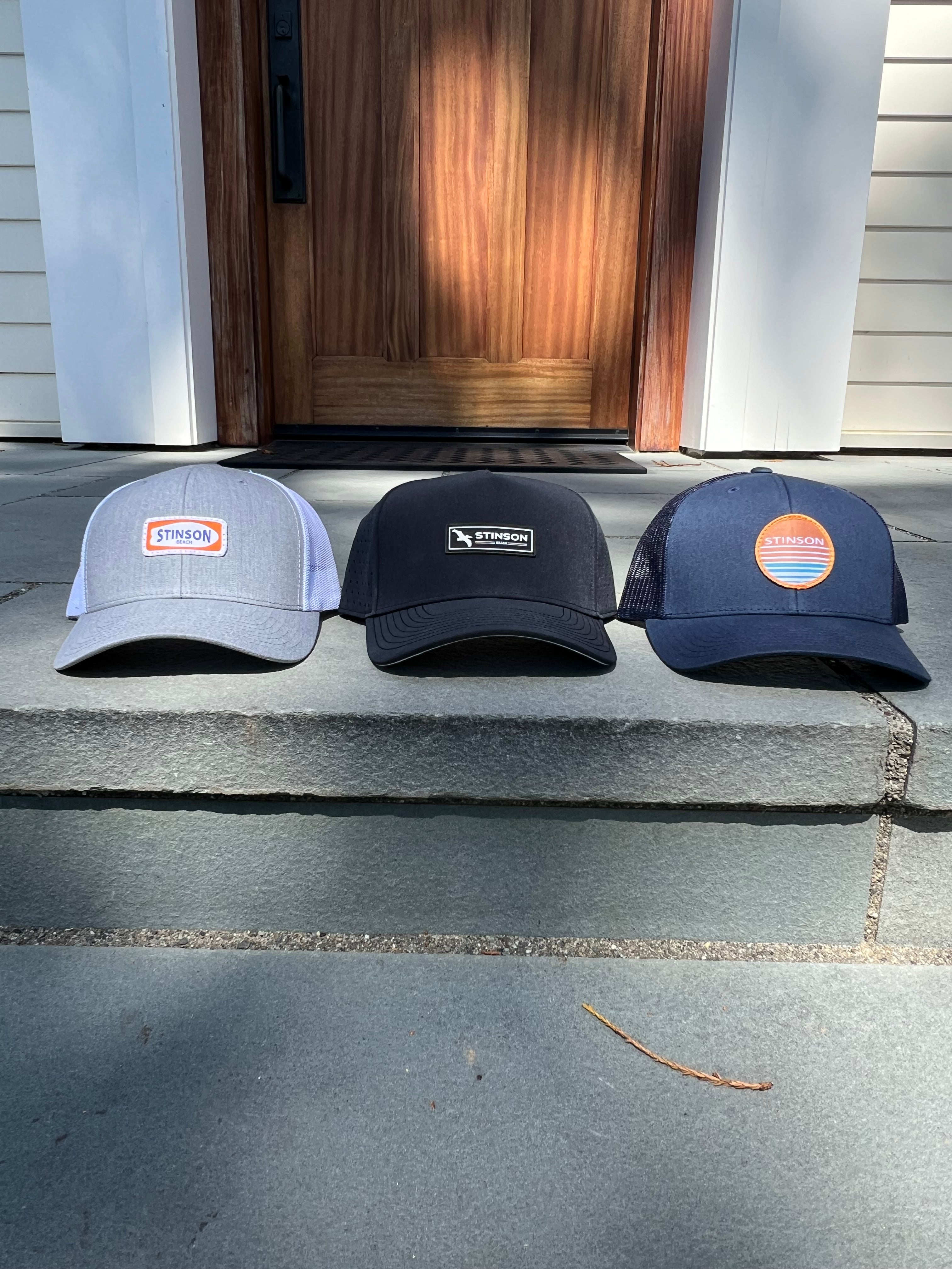 Three trucker-style baseball caps are lined up on concrete steps in front of a wooden front door. The left cap is light gray with an orange and white patch; the center cap is black with a black rectangular patch; the right cap is navy blue with a circular orange and blue patch.