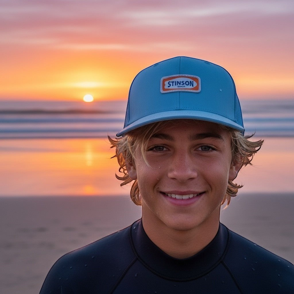 Lowkey Local Stinson Beach trucker hat in Carolina blue with Stinson Beach patch in white and orange worn by a youngster at sunset on the beach.