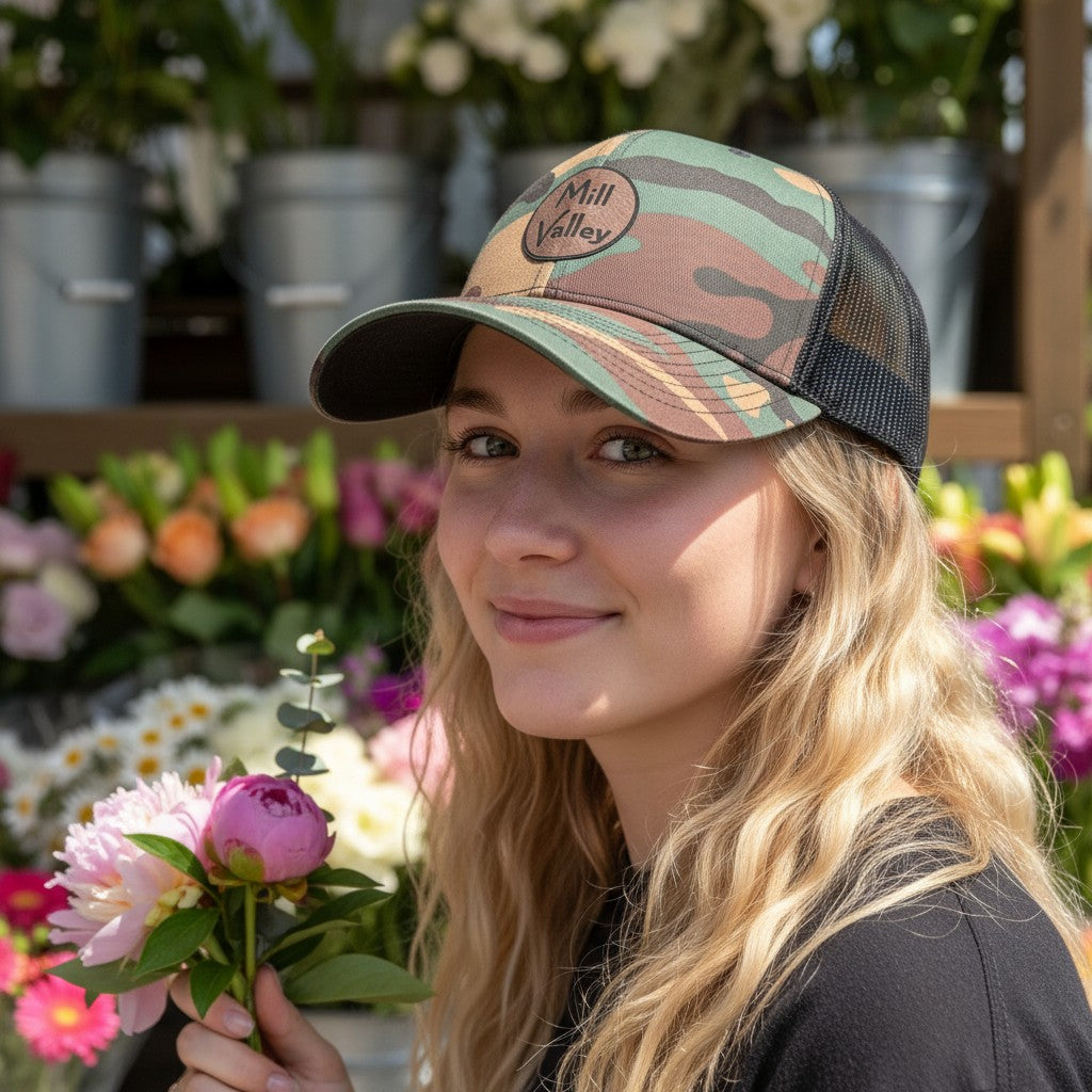 Lowkey Local Mill Valley camo trucker hat with black mesh back and circular logo patch worn by a woman in a flower shop