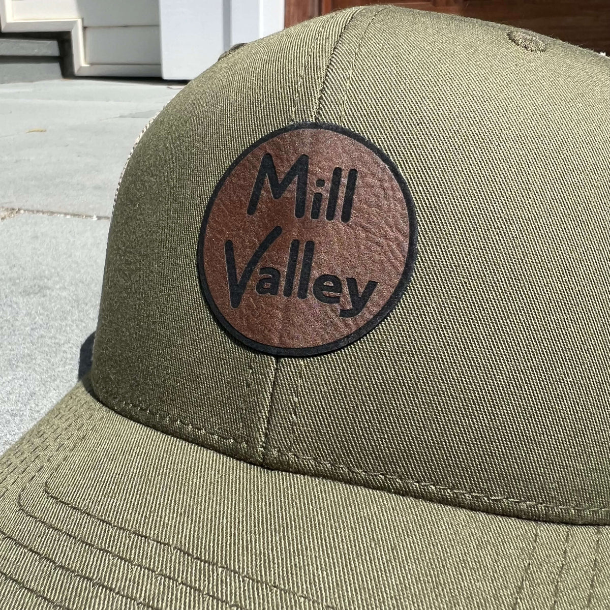 Lowkey Local moss green structured trucker hat with a circular brown leather Mill Valley logo patch on the front.