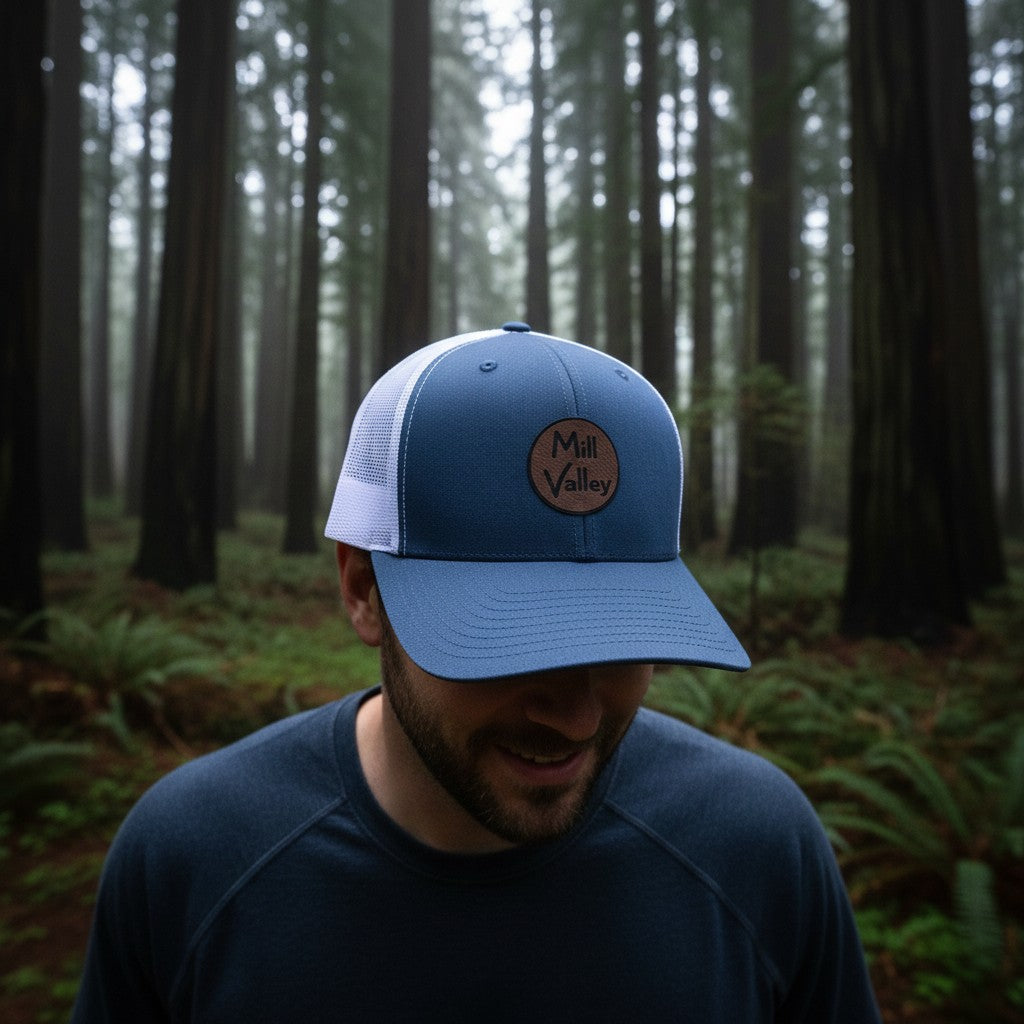 Lowkey Local navy blue trucker hat with a circular Mill Valley patch worn by a man in a redwood forest.