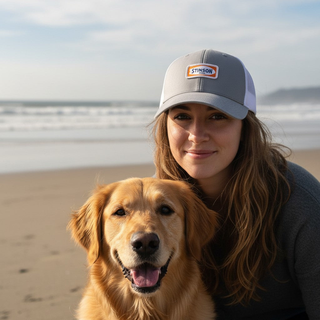 Lowkey Local Stinson Beach trucker hat in grey and white mesh worn by a woman with a golden retriever on the beach.