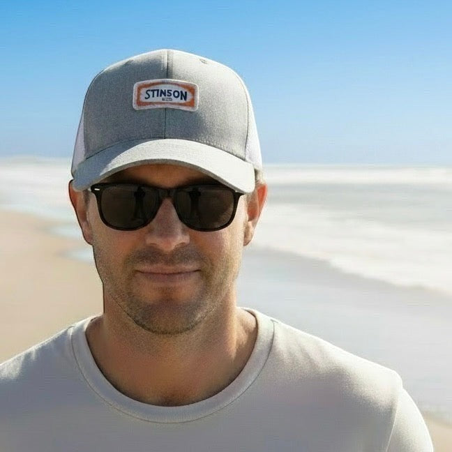 Man wearing a Lowkey Local grey and white mesh trucker hat with a Stinson Beach embroidered patch on a sunny beach.