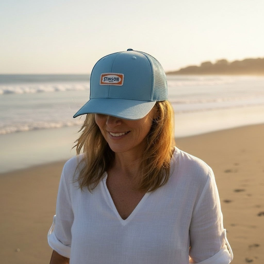 Lowkey Local Stinson Beach trucker hat in Carolina blue with a surf-themed patch worn by a woman on the beach.