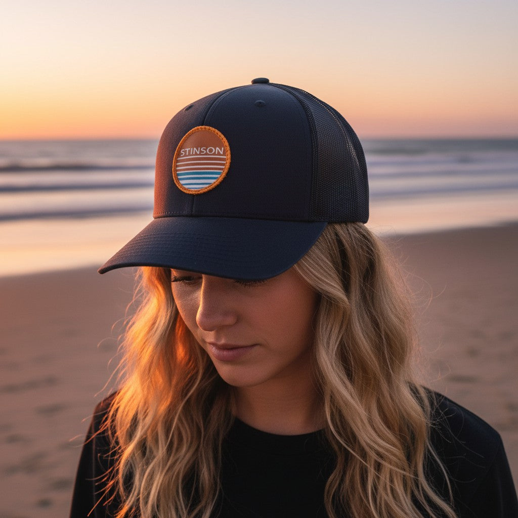 Lowkey Local Stinson Sunset navy trucker hat with a circular patch worn by a woman on a beach at sunset
