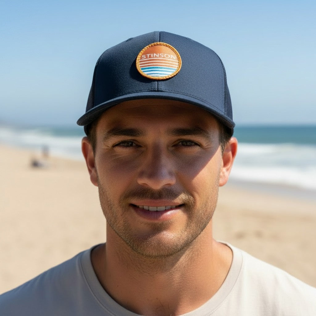 Lowkey Local Stinson Sunset navy trucker hat with a circular stitched patch worn by a man on a sunny beach